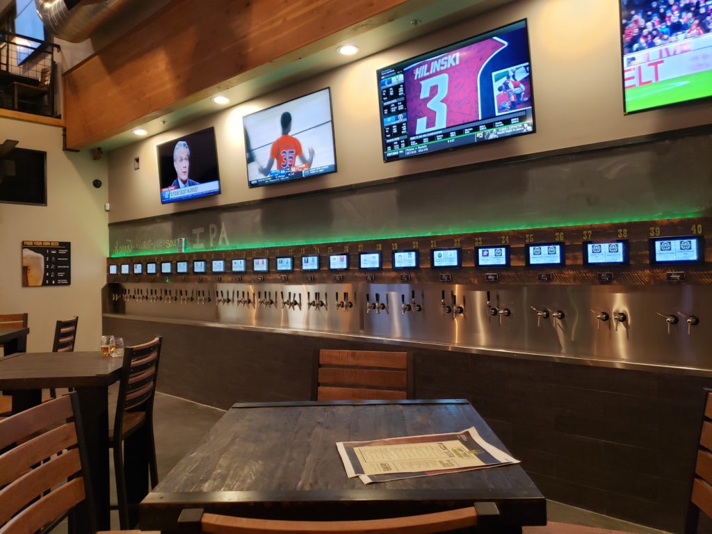 A self-pour drink wall in a restaurant with three tv screens above