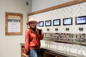 A smiling woman stands in front of a PourMyBeer tap wall with a full glass.