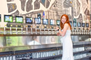 A woman smiling and pointing at self-pour beer taps inside a restaurant