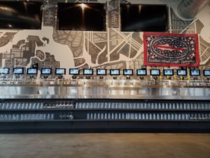 A self-pour beer wall with multiple taps, shelves of organized glasses, and TV screens above the taps.